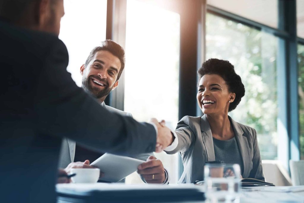 An image of three people sitting at a table. Two are facing the camera, one has got their back to it. The person with their back to the camera is shaking hands with one of the others.